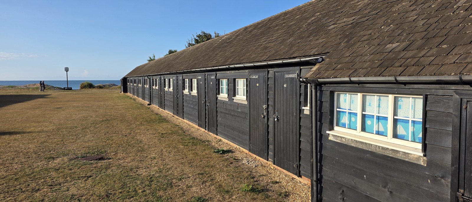 A long terrace of black timber beach huts with a tiled roof, numbered doors and white window frames, one window curtained in a blue beach hut print, with the Greensward and the sea beyond
