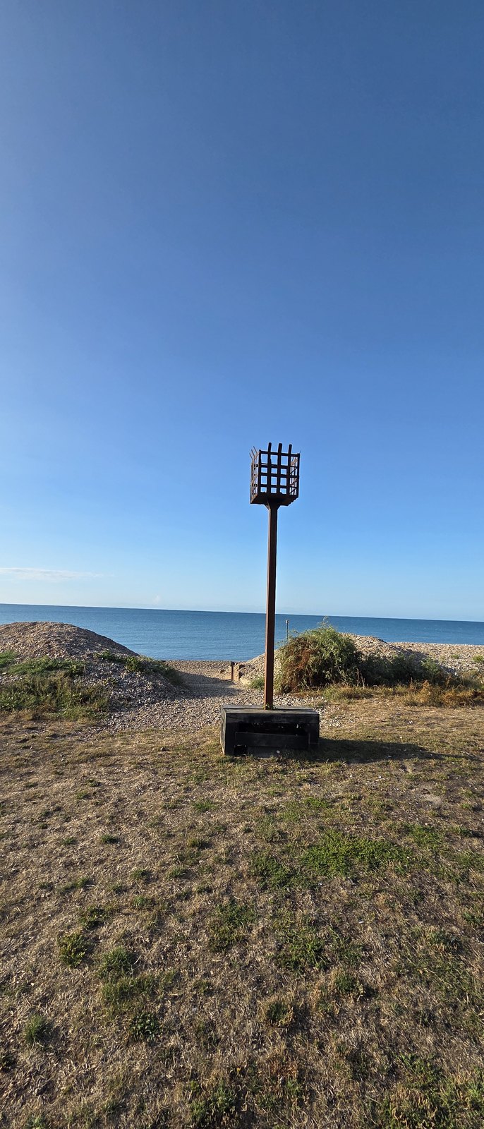 A tall wooden beacon post topped with an iron fire basket, standing on the vegetated shingle of the Greensward with the sea and horizon beyond