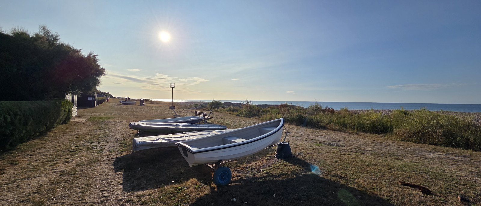 Several small boats under covers on trailers on the mown grass of the Greensward, with the beacon, the shingle bank and the sea beyond in low evening sun