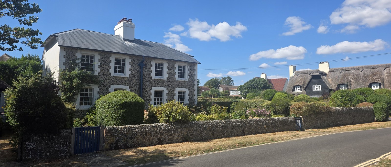 A two-storey house faced with knapped flint, with painted brick quoins and window surrounds, sash windows, a slate roof and a white chimney, behind a low flint wall with a blue gate, and a thatched cottage beyond