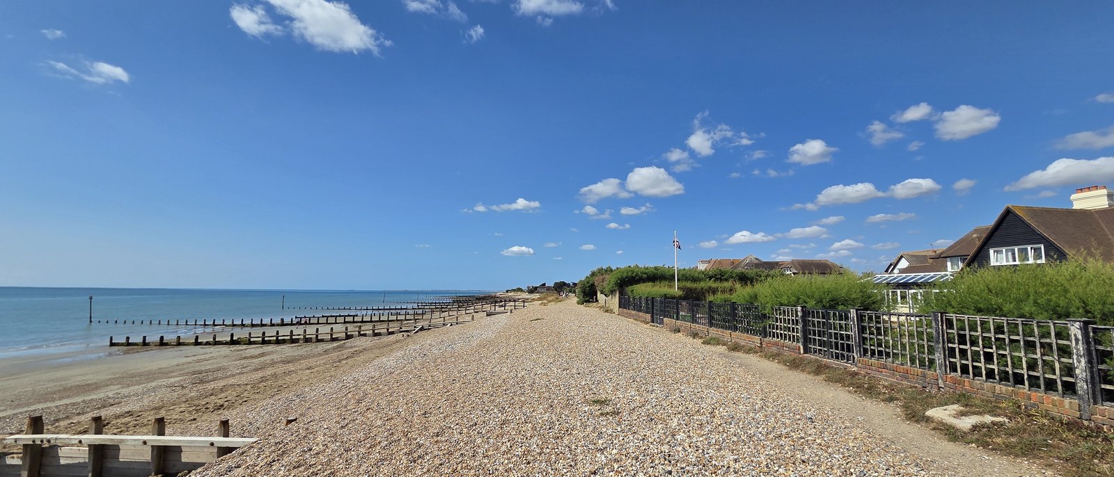 Looking west along the shingle bank of the Greensward, with timber groynes running out into a calm sea on the left, fences and houses on the right, a flagpole flying a Union flag, and a wide blue sky