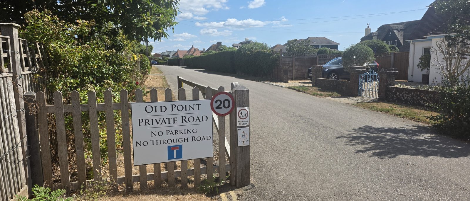 A sign at the entrance to Old Point reading private road, no parking, no through road, beside a 20 mph roundel and smaller signs asking people to keep dogs on leads and clean up after them, with the unadopted road running away between grass verges