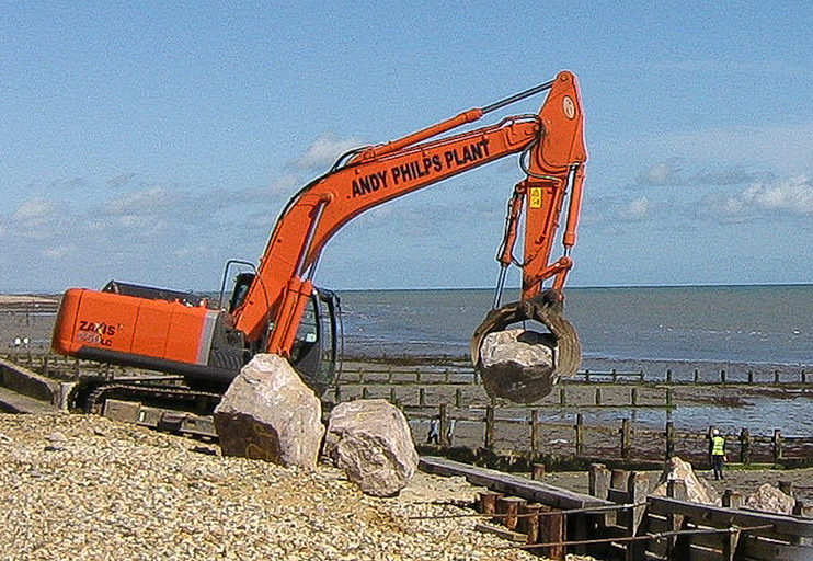 A tracked crane working on the beach placing rock armour beside a timber groyne