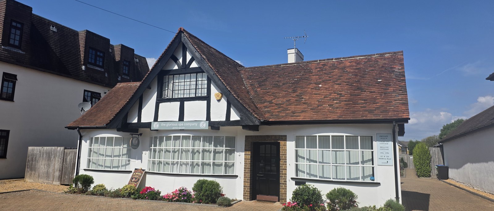 A white and black timbered cottage with a steep tiled roof and bay windows, signed the glorious spa company, with flower beds along the front