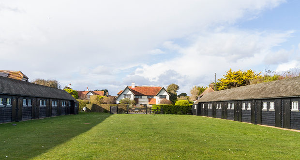 Two rows of black timber beach huts across mown grass on the Greensward