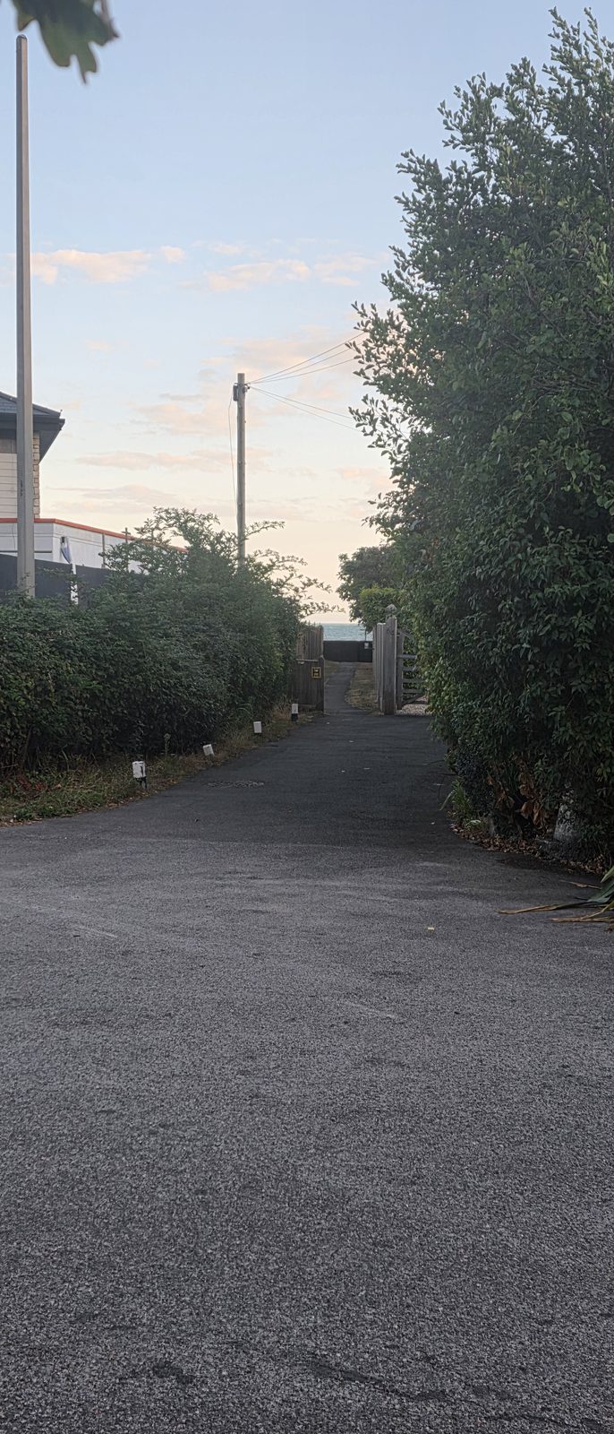 A narrow tarmac path running between hedges and a wooden gate, heading towards the sea under an evening sky